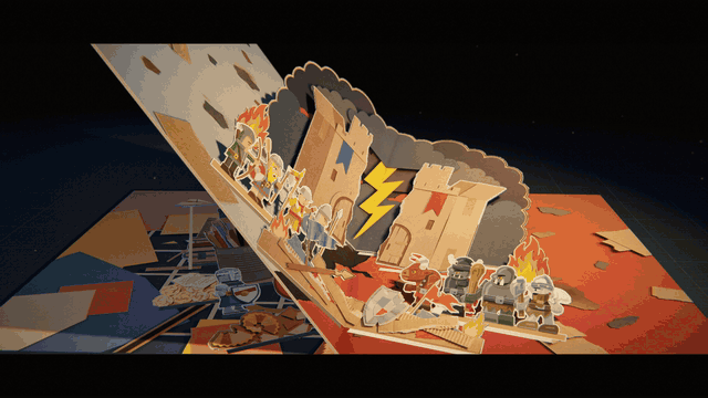 The second spread of the pop-up book under the desk lamp: two cardboard castles fold up out of the page with a yellow lightning bolt between them, a blue army in front of one and a red army in front of the other, while the caption under it unfolds, 'From corrugated walls, a new war is unfolding.'